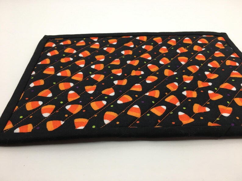 May include: A black quilted placemat featuring a pattern of orange, white, and yellow candy corn. The placemat has a black border and is decorated with small dots of green, purple, and yellow.
