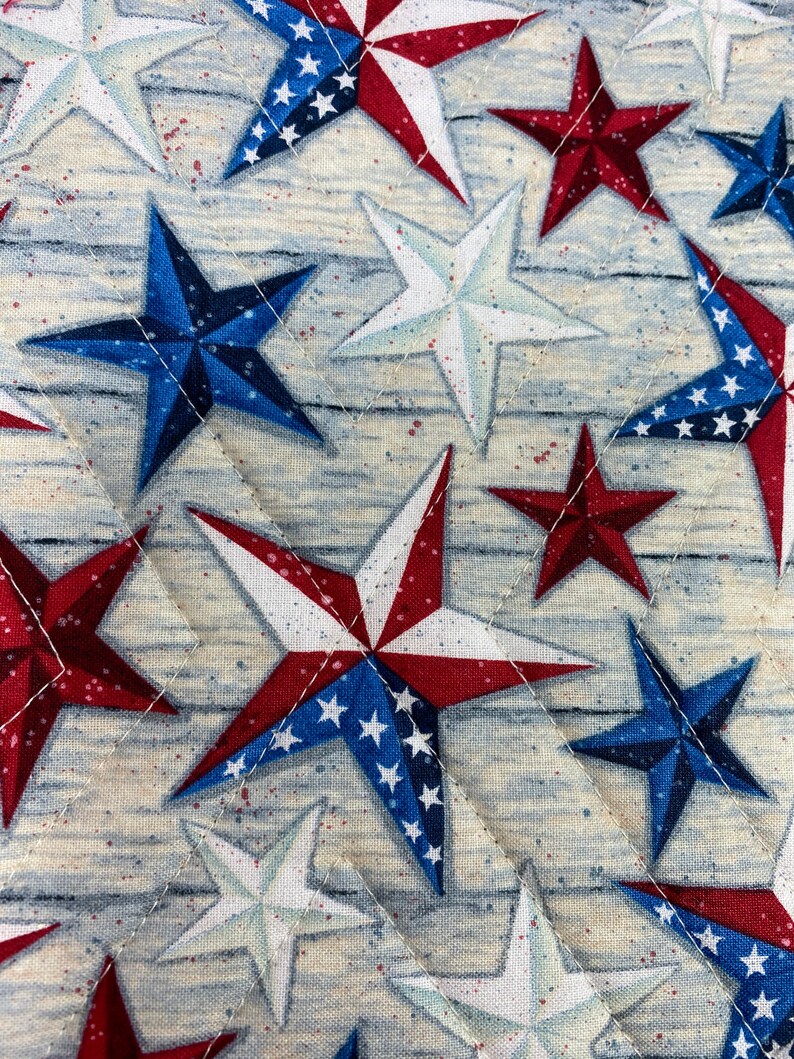 May include: A quilted fabric with a repeating pattern of red, white, and blue stars. The stars are in various sizes and orientations, and some have a white background with blue stars.