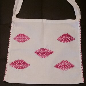 May include: White cotton tote bag with a pink diamond pattern embroidered on the front. The bag has a single strap and a zig-zag stitch around the edges.