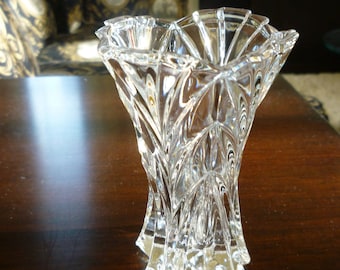 May include: A clear glass vase featuring a multifaceted, geometric design with vertical ridges and a scalloped, flared rim. The body of the vase has a tapered shape that widens toward the top, creating a series of reflective, angular surfaces.