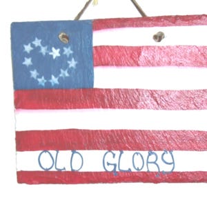 Puede incluir: Una placa rectangular de pizarra pintada con el diseño de la bandera estadounidense, con franjas horizontales rojas y blancas y un cantón azul con estrellas blancas dispuestas en forma de corazón. El texto OLD GLORY está pintado en azul.