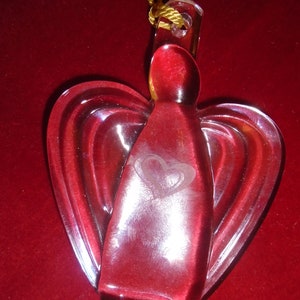 Puede incluir: Un adorno de cristal rojo intenso translúcido con forma de ángel y alas estilizadas. El cuerpo central presenta un diseño de corazón grabado. Un cordón dorado pasa por un orificio en la parte superior para colgarlo y dejar pasar la luz.