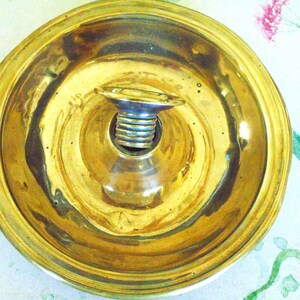 May include: A circular brass candle holder base with a polished gold-toned finish. The center features a raised, threaded silver-toned metal socket designed to secure a candle. The surface of the brass is smooth and reflective with a wide, flat rim.