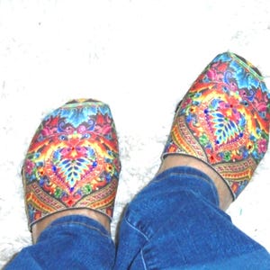 Puede incluir: Un par de zapatos sin cordones con un vibrante estampado floral y de cachemira multicolor con pequeños diamantes de imitación incrustados, usados con jeans azules. El calzado tiene un diseño de punta redonda y una silueta baja de tela.