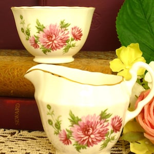Puede incluir: Un juego de lechera y azucarero de porcelana color crema con un delicado diseño floral de crisantemos rosas, hojas verdes y bordes festoneados dorados. Las piezas tienen una base redondeada con pie y un asa curva en la lechera.