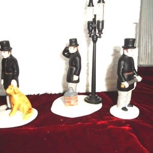 May include: A collection of miniature figurines depicting Victorian-era figures. The set includes a man with a dog, a man saluting, and a man with a rolled paper. A black lamppost and a small building are also included. The figurines are on white bases.