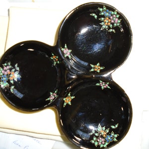 Puede incluir: Un plato de servicio negro de tres secciones con acabado brillante. Cada uno de los tres cuencos circulares está decorado con pequeños racimos de flores coloridas en tonos rosa, azul, naranja y verde, con delicados contornos metálicos.