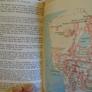 May include: A vintage map of the ancient Greek island of Delos, showing the location of the Sacred Lake, the Agora of the Compitaliasts, the Theatre, the Sanctuary of Apollo, and other important sites. The map is printed in red and black on a cream-colored background.