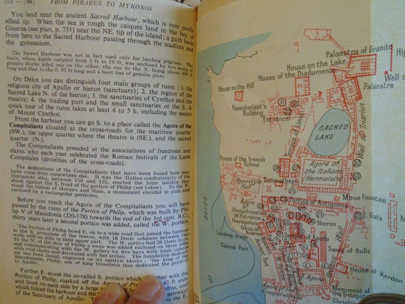 May include: A vintage map of the ancient Greek island of Delos, showing the location of the Sacred Lake, the Agora of the Compitaliasts, the Theatre, the Sanctuary of Apollo, and other important sites. The map is printed in red and black on a cream-colored background.