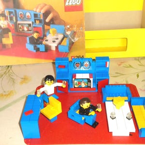 May include: A LEGO set 264 living room scene featuring a red baseplate, blue furniture, a white table, yellow chairs, and two minifigures. The set includes a blue wall unit with a television screen graphic and a box with the LEGO logo.