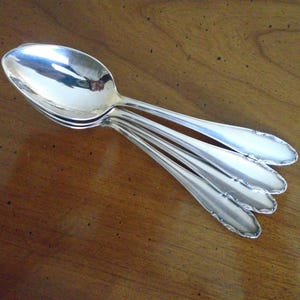 May include: A set of four silver spoons arranged on a wooden surface. The spoons have a reflective, polished finish, with a decorative edge along the spoon bowls. The spoons are angled, showcasing their design and the wood grain.