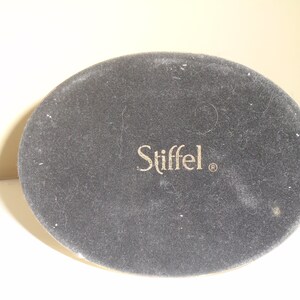 Puede incluir: Primer plano de la base de un objeto ovalado gris oscuro con la palabra "Stiffel" en letras doradas. El objeto tiene una textura similar al fieltro.