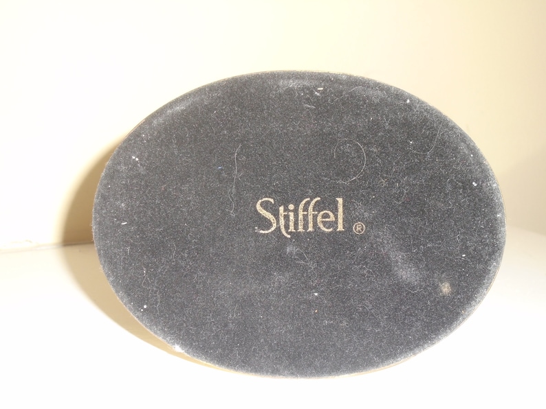 Puede incluir: Primer plano de la base de un objeto ovalado gris oscuro con la palabra "Stiffel" en letras doradas. El objeto tiene una textura similar al fieltro.