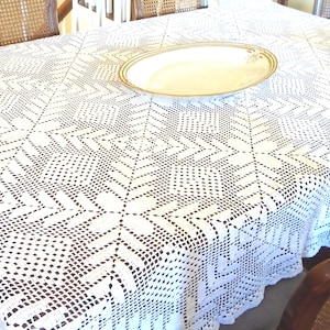 Puede incluir: Un mantel blanco, intrincadamente tejido a ganchillo, con un patrón geométrico, cubre una mesa. Una fuente ovalada con borde dorado está sobre la mesa. El diseño del mantel presenta un motivo de estrella repetido.