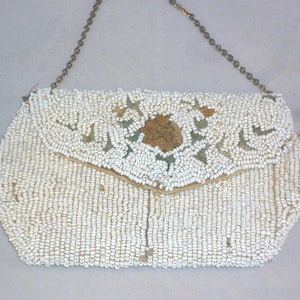 Puede incluir: Un bolso de mano tipo sobre cubierto de cuentas blancas con una correa de cadena metálica dorada. El exterior está recubierto de pequeñas cuentas blancas con un motivo floral central en tonos dorados y verdes en la solapa puntiaguda.