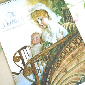 Puede incluir: Un calendario de compromisos de 1984 titulado The Dollhouse con una imagen impresa de dos muñecas antiguas con gorros y ropa blanca sentadas en un cochecito de mimbre marrón. El calendario tiene una espiral en la parte superior.