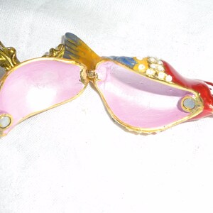 May include: A red, yellow, and blue bird trinket box with a pink interior and a gold leaf design. The box is open and the lid is hinged.