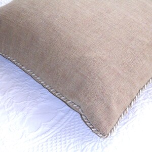 Puede incluir: Un cojín rectangular beige con una superficie texturizada y un ribete de cuerda plateada. El cojín está sobre una superficie blanca acolchada.