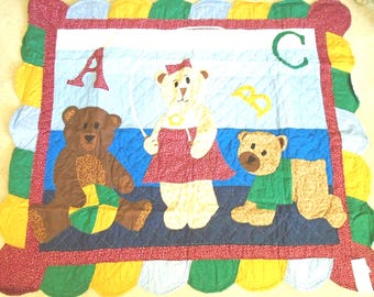 Puede incluir: Una manta acolchada con tres osos de peluche y las letras A, B y C. El oso central sostiene una cuerda de saltar, el oso de la izquierda está sentado con una pelota y el de la derecha gatea. El borde tiene festones rojos, amarillos, verdes y azules.