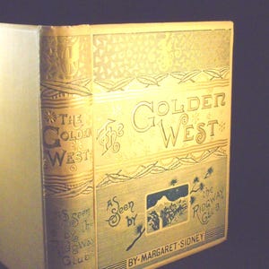 Puede incluir: Un libro antiguo titulado "The Golden West" con letras doradas y detalles decorativos. La portada presenta el texto "As Seen by the Ridgway Club" y el nombre del autor, Margaret Sidney. El libro tiene un lomo de color crema.