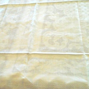 Puede incluir: Un mantel rectangular de color crema que presenta un intrincado patrón de rejilla calada combinado con secciones tejidas sólidas que forman grandes motivos florales y de volutas repetitivos en toda la superficie de la tela.