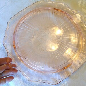 Puede incluir: Un plato de vidrio rosa con borde festoneado. El plato tiene un dise&ntilde;o floral en el centro y l&iacute;neas radiales. El plato est&aacute; sostenido por una mano.