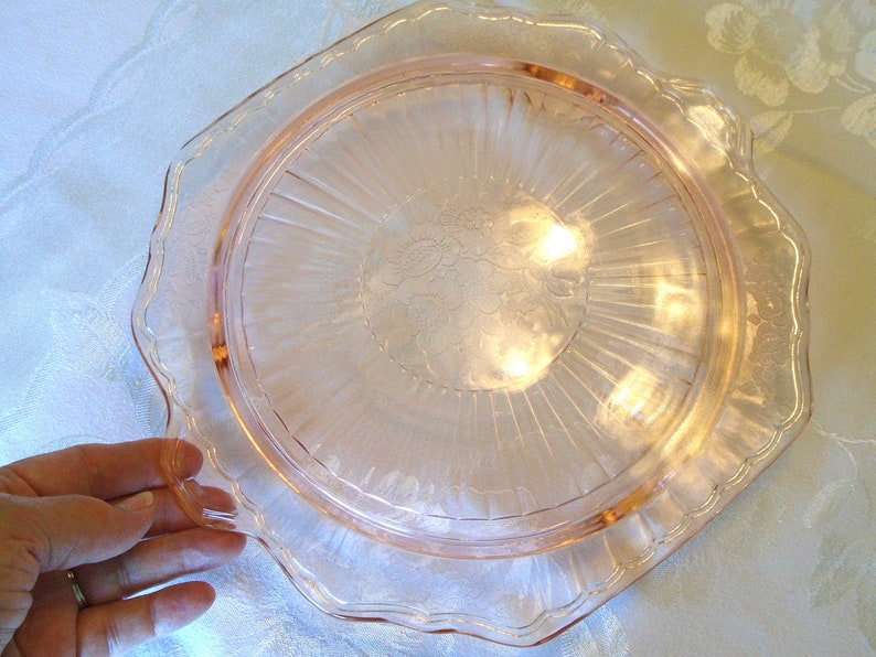 Puede incluir: Un plato de vidrio rosa con borde festoneado. El plato tiene un dise&ntilde;o floral en el centro y l&iacute;neas radiales. El plato est&aacute; sostenido por una mano.