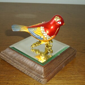 May include: A small, red, yellow, and blue bird figurine with rhinestones on a mirrored base. The bird is perched on a gold-colored branch.