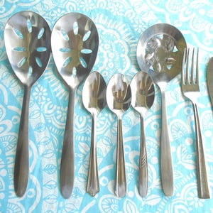 May include: A collection of eight stainless steel utensils including two large slotted serving spoons, three small teaspoons, one round slotted serving spoon with a floral pattern, one dinner fork, and one dinner knife with a smooth finish.