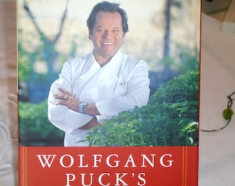 May include: A book cover featuring a portrait of Wolfgang Puck, a famous chef, in a white chef's jacket. He is standing in front of a green bush and smiling. The book title is "Wolfgang Puck's" in white text on a red background.