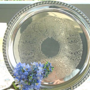 Puede incluir: Una bandeja de servicio redonda de tono plateado con un intrincado patrón floral y de volutas en relieve en el centro y un borde de estilo cuerda trenzada en el exterior. Un grupo de pequeñas flores azules se sitúa en primer plano.