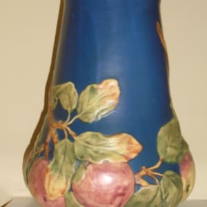 May include: A blue ceramic vase with a wide mouth and a narrow base. The vase is decorated with a raised design of apples and leaves in shades of green, yellow, and pink.