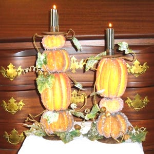 Op de afbeelding: Twee decoratieve kandelaars met gestapelde pompoenontwerpen. Elke houder heeft drie oranje pompoenen, groene bladeren en een brandende kaars bovenop. De houders staan op een wit oppervlak tegen een donkere houten achtergrond.
