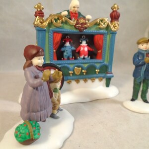 Puede incluir: Un puesto de títeres de madera de estilo vintage con una cortina roja, detalles dorados y dos títeres. Dos niños están viendo el espectáculo, y un hombre está operando los títeres desde arriba. La escena está ambientada en un país de las maravillas invernal con nieve en el suelo.