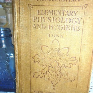 Puede incluir: Un libro antiguo titulado "Elementary Physiology and Hygiene" con "Indiana Edition" en la portada. La portada es de color beige con texto en relieve y un diseño decorativo de hojas y bellotas. Una botella de vidrio azul está al lado del libro.