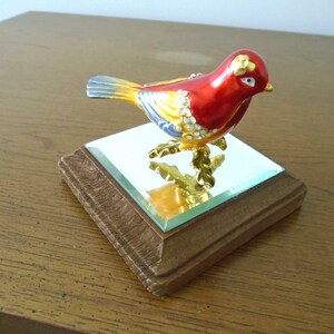 May include: A small, red, yellow, and blue bird figurine with gold accents and rhinestones. The bird is perched on a small, square, wooden base with a mirrored top.