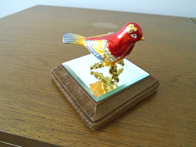 May include: A small, red, yellow, and blue bird figurine with gold accents and rhinestones. The bird is perched on a small, square, wooden base with a mirrored top.