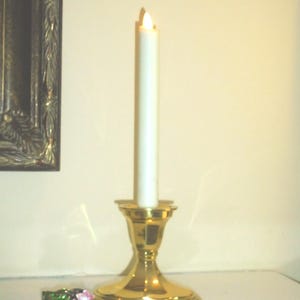Puede incluir: Una vela encendida de color blanquecino en un candelabro dorado. El candelabro tiene una base ancha y una parte superior acampanada. Un peque&ntilde;o objeto decorativo enjoyado con detalles rosas y verdes est&aacute; cerca. El fondo es de color neutro.