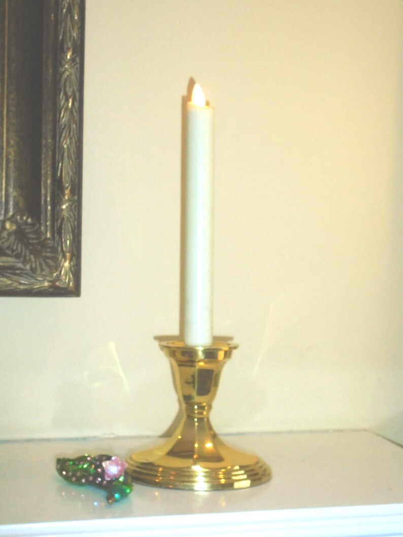 Puede incluir: Una vela encendida de color blanquecino en un candelabro dorado. El candelabro tiene una base ancha y una parte superior acampanada. Un peque&ntilde;o objeto decorativo enjoyado con detalles rosas y verdes est&aacute; cerca. El fondo es de color neutro.
