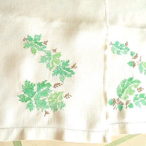Puede incluir: Tela blanquecina con dise&ntilde;os de hojas bordadas a punto de cruz en verde y marr&oacute;n. Las hojas son de diferentes tonos de verde, con tallos y detalles marrones. La tela tiene una textura sutil y un dobladillo cosido.