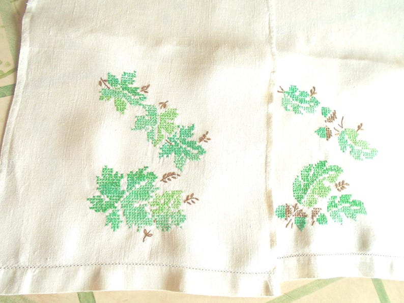 Puede incluir: Tela blanquecina con dise&ntilde;os de hojas bordadas a punto de cruz en verde y marr&oacute;n. Las hojas son de diferentes tonos de verde, con tallos y detalles marrones. La tela tiene una textura sutil y un dobladillo cosido.