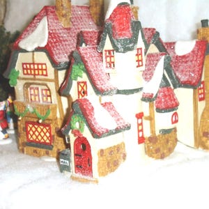 Può includere: Una casa in ceramica di un villaggio natalizio con tetti rossi spolverati di neve. La casa ha più livelli, finestre e una porta rossa con una cassetta delle lettere. La vegetazione decorativa e le superfici innevate aggiungono alla scena festosa.