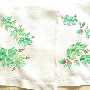 Puede incluir: Dos servilletas de lino blanco roto con dise&ntilde;os de hojas bordadas en verde y marr&oacute;n. Las hojas son de diferentes tonos de verde, y los tallos y acentos son marrones. Las servilletas tienen un dobladillo simple cosido.