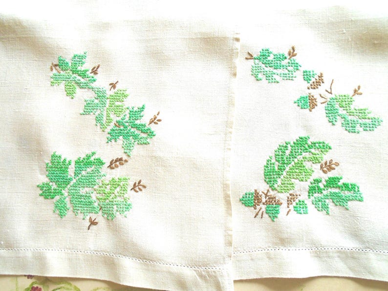 Puede incluir: Dos servilletas de lino blanco roto con dise&ntilde;os de hojas bordadas en verde y marr&oacute;n. Las hojas son de diferentes tonos de verde, y los tallos y acentos son marrones. Las servilletas tienen un dobladillo simple cosido.