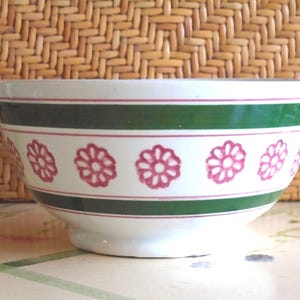 May include: White ceramic mixing bowl with a green and red floral design. The bowl features a wide green band with red stripes and pink floral patterns. The bowl has a pedestal base and is set against a woven background.