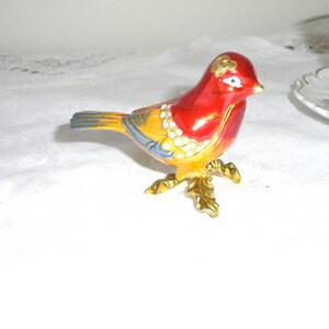 May include: A small, red, yellow, and blue bird figurine with gold accents and rhinestones. The bird is perched on a gold stand with leaves.