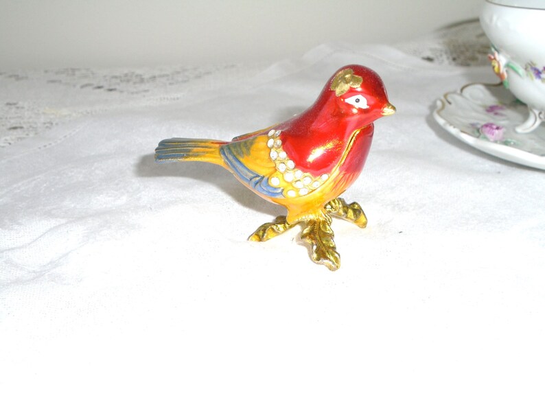 May include: A small, red, yellow, and blue bird figurine with gold accents and rhinestones. The bird is perched on a gold stand with leaves.