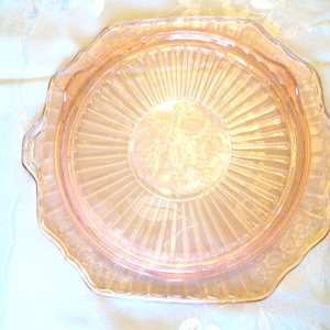 Puede incluir: Una fuente de vidrio color melocot&oacute;n vintage con borde festoneado y dos asas. La fuente presenta un dise&ntilde;o radial con un motivo floral en el centro. La fuente tiene aproximadamente 25 cm de di&aacute;metro.