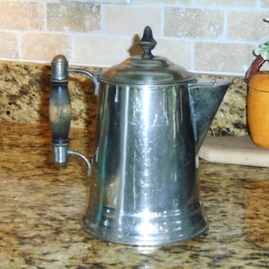 May include: A vintage silver coffee pot with a dark wooden handle and a lid. The pot has a classic design with a spout for pouring. The handle is attached to the side of the pot. The pot is sitting on a countertop.