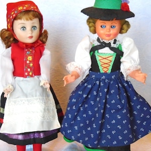 Puede incluir: Dos muñecas vintage vestidas con trajes folclóricos europeos tradicionales. Una muñeca lleva un chaleco rojo con detalles florales, un delantal blanco y un gorro rojo. La otra muñeca lleva una falda azul estampada, un corpiño negro con cordones naranjas y un sombrero verde.